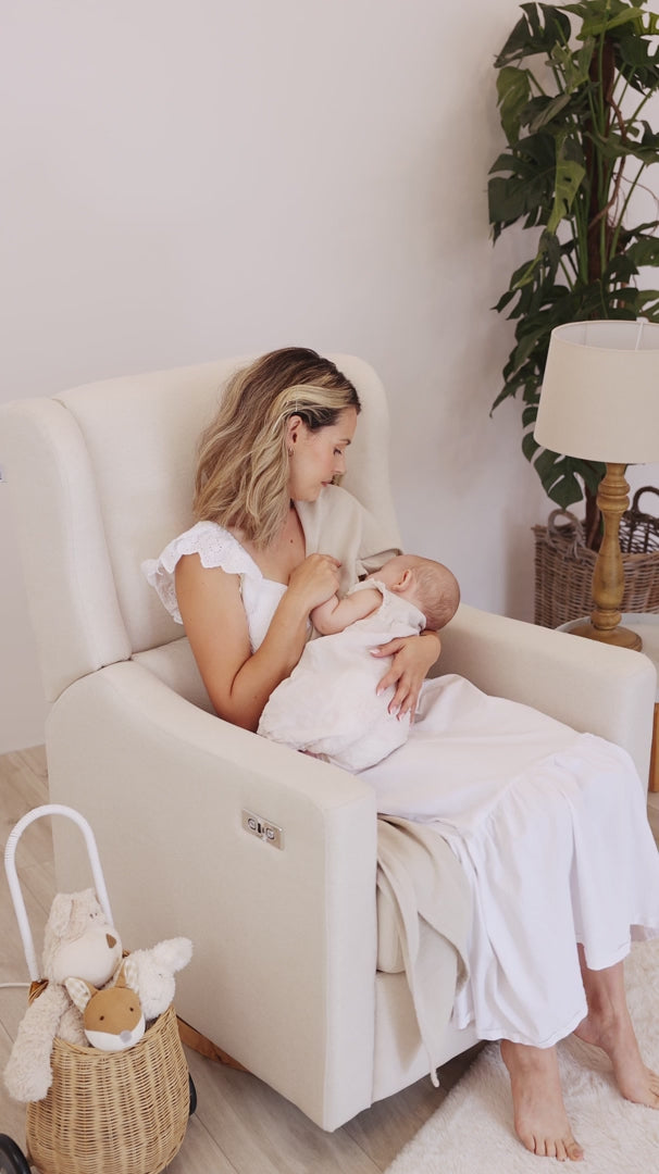 Mother cradling her baby in the Gaia Baby Lyra nursing chair in Oat beside a wicker toy pram